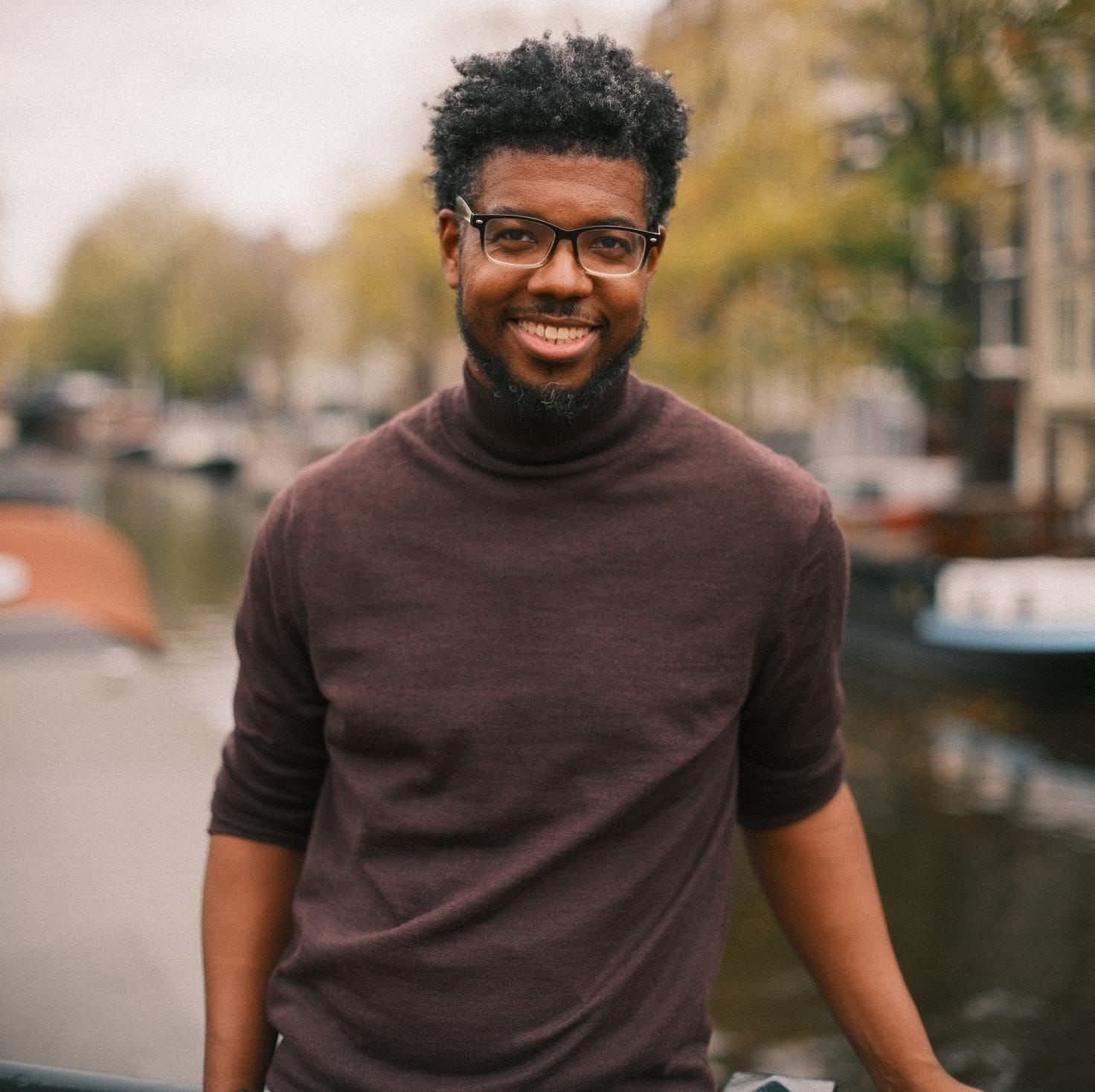 Portrait of Atnre Alleyne smiling, wearing glasses and a brown turtleneck